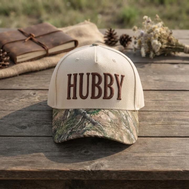 Close-up of cream Hubby trucker hat with raised brown lettering and camo bill