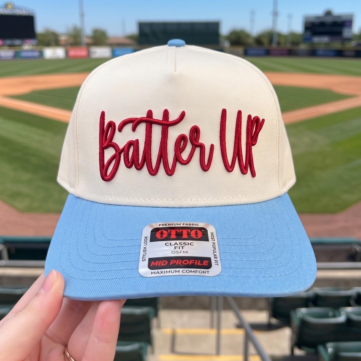 Baseball cap with embroidered 'Batter Up' design in cream and light blue displayed in a stadium setting
