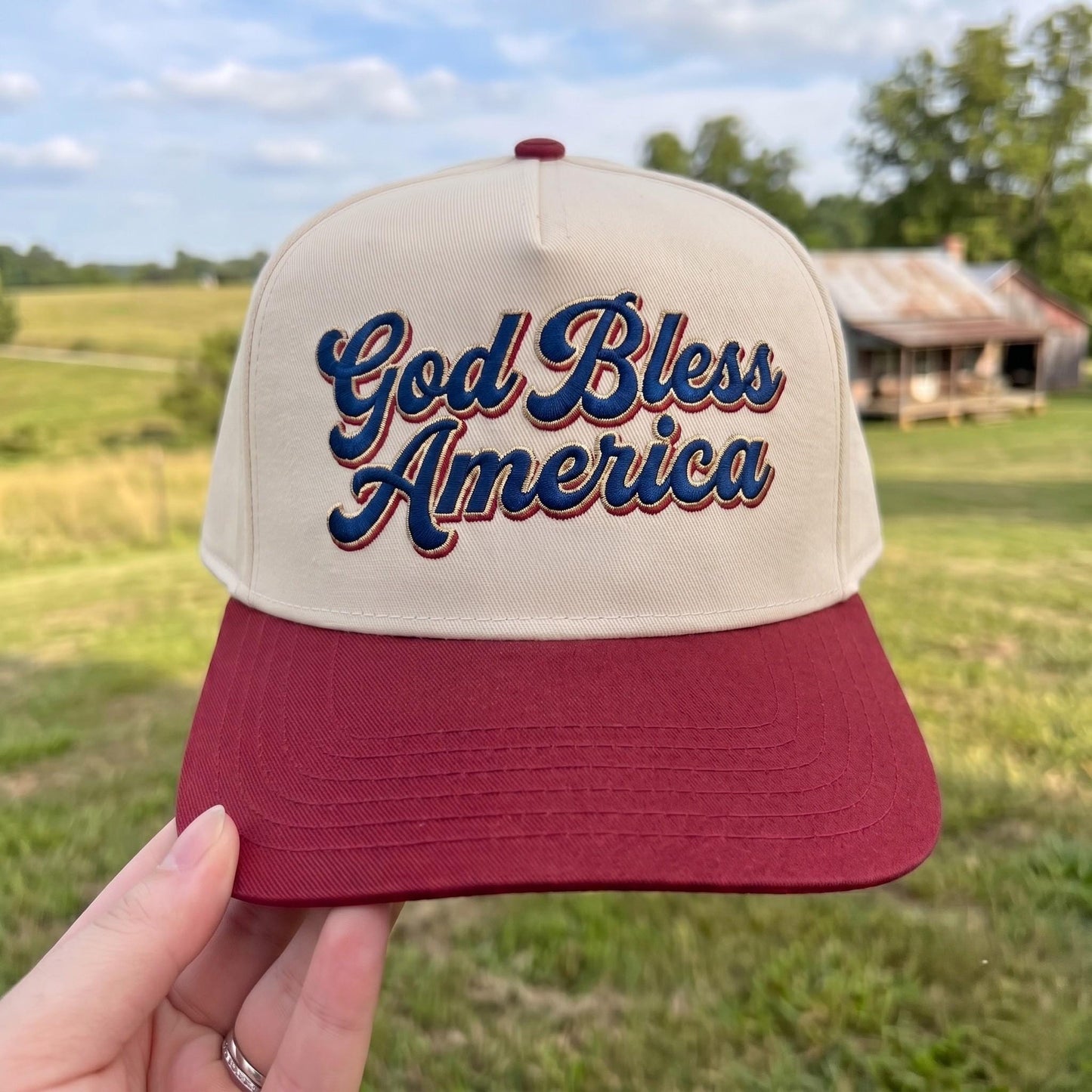 Patriotic trucker hat with 'God Bless America' embroidery in red and beige on a grassy field