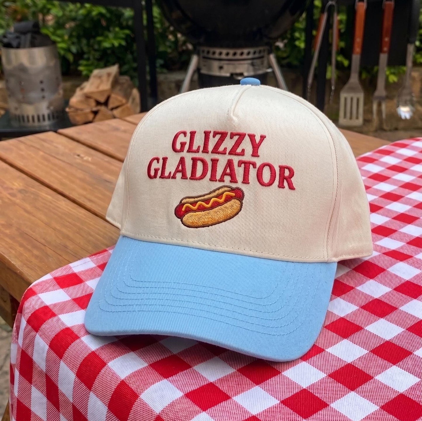 Novelty cap with 'Glizzy Gladiator' embroidery in cream with blue bill on picnic table