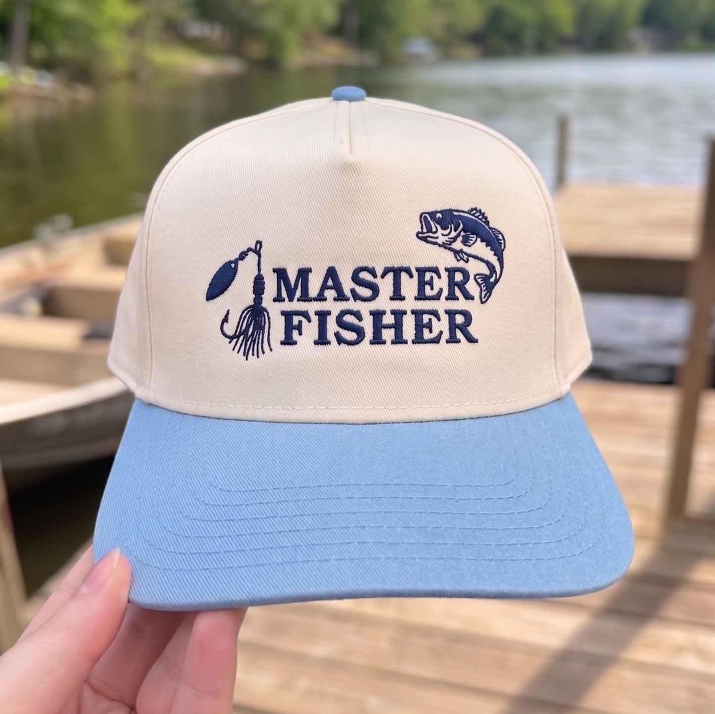 Fishing cap with beige crown and blue brim, embroidered with 'Master Fisher', displayed near a lake