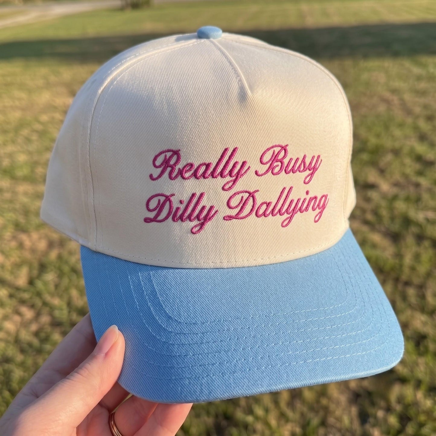 Embroidered baseball hat with 'Really Busy Dilly Dallying' in pink text on cream with light blue brim