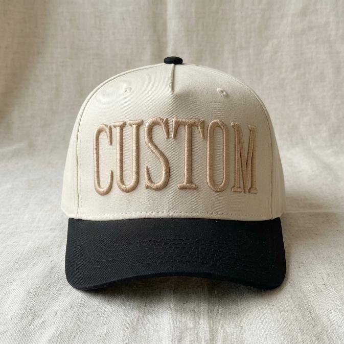 Front view of a cream and black two-tone trucker hat with embroidered CUSTOM lettering