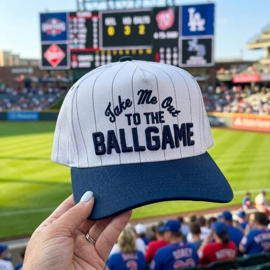 White pinstripe baseball hat with navy brim and Take Me Out to the Ballgame embroidery at a baseball stadium