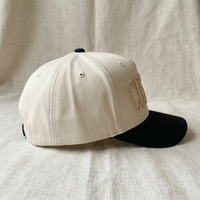 Side view of the cream trucker-style hat showing the black brim and front embroidery