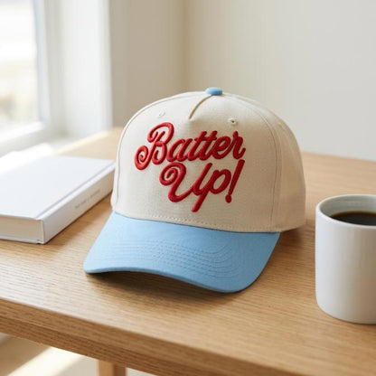 Two-tone Batter Up trucker cap with red embroidered script, cream front, and light blue brim on a wooden table