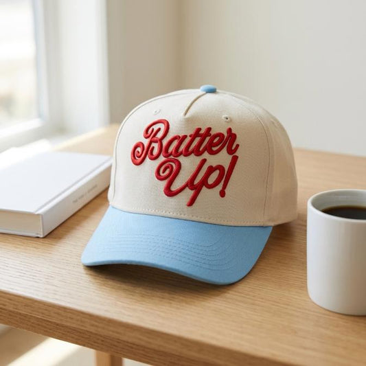 Two-tone Batter Up trucker cap with red embroidered script, cream front, and light blue brim on a wooden table