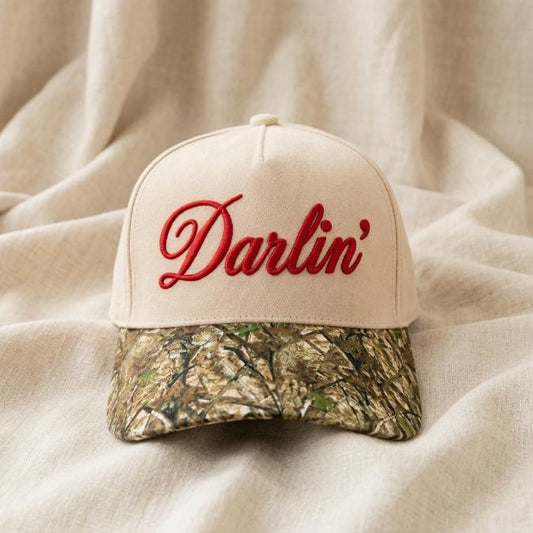 Front view of a neutral Darlin' embroidered hat with red script lettering and camouflage brim