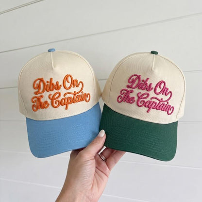 Two cream two-tone trucker caps embroidered with Dibs On The Captain, shown with blue and forest green bills
