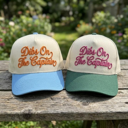 Close-up of Dibs On The Captain embroidered trucker caps displayed outdoors on a wooden table
