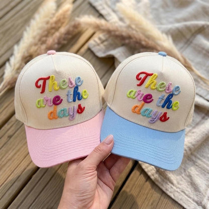 Two two-tone embroidered hats with colorful These are the days lettering, shown in pink and light blue