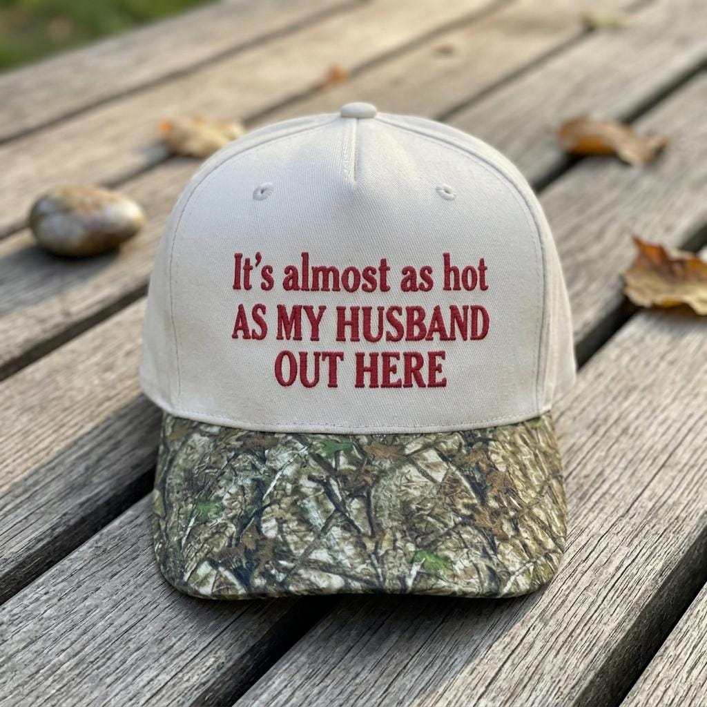 White hat with camouflage brim and red text reading “It’s almost as hot as my husband out there”