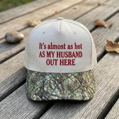 White hat with camouflage brim and red text reading “It’s almost as hot as my husband out there”