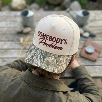 Person wearing the Somebody’s Problem hat with cream crown and camouflage bill