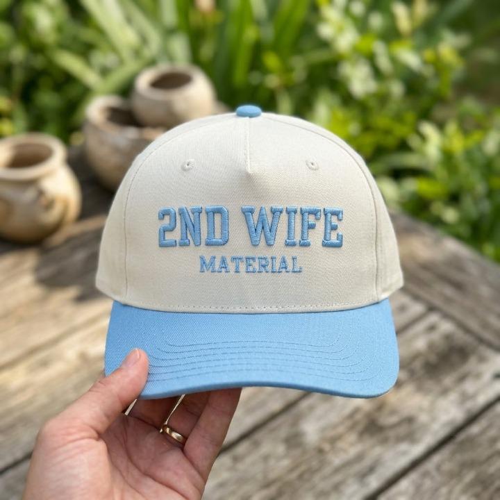Cream and light blue embroidered 2ND WIFE MATERIAL hat held outdoors