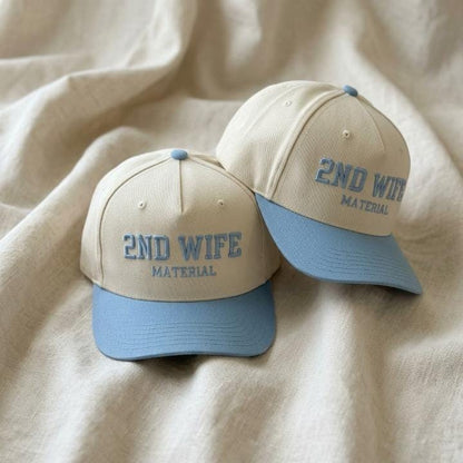 Two cream and light blue 2ND WIFE MATERIAL embroidered trucker hats on a soft fabric background