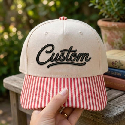 Cream embroidered hat with black Custom script and red-and-white striped bill outdoors