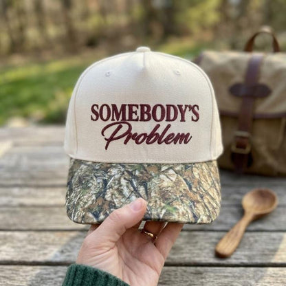 Front view of cream embroidered hat with Somebody’s Problem burgundy lettering and camouflage-patterned bill