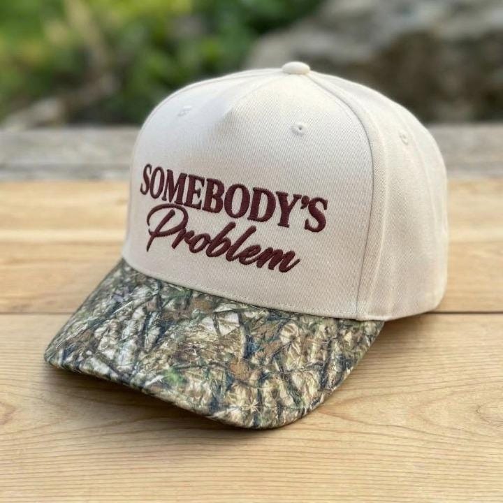 Angled view of embroidered Somebody’s Problem hat with camouflage bill