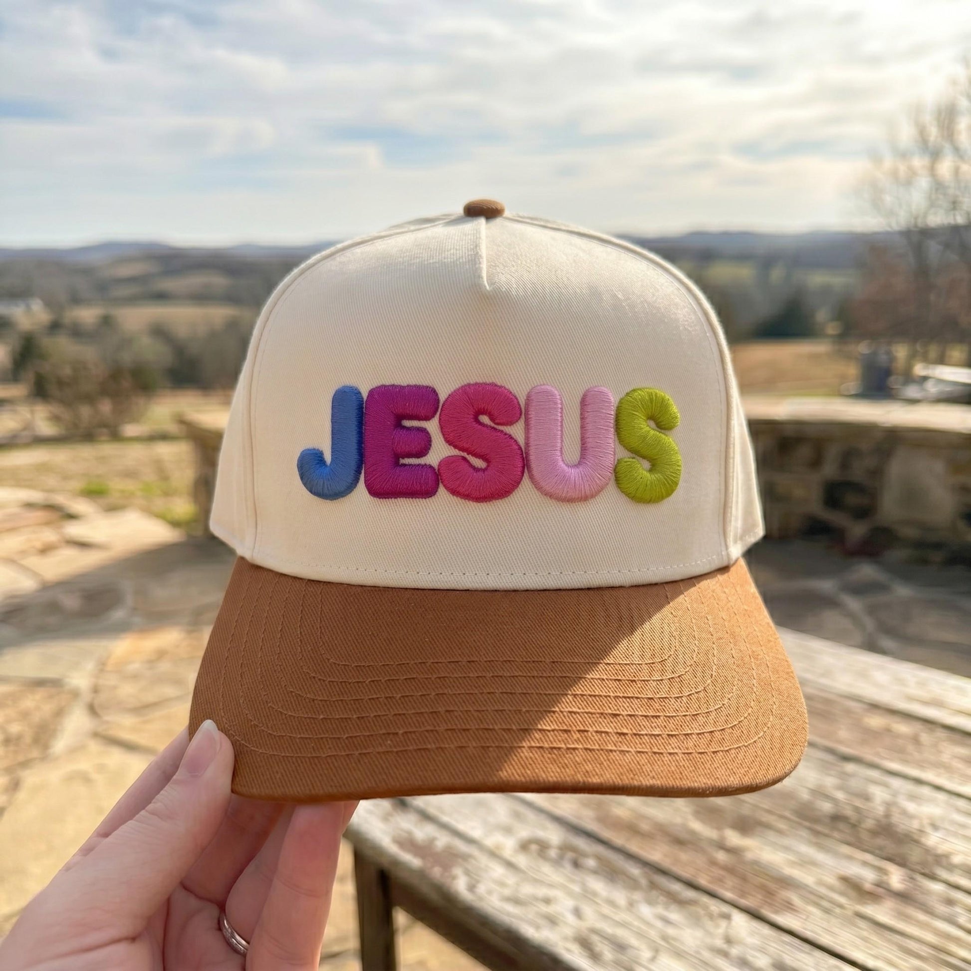 Colorful embroidered Jesus message cap with multicolor letters and a brown brim displayed outdoors