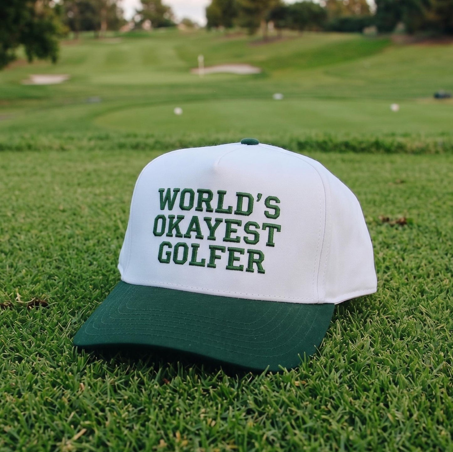 Novelty golf cap with 'World's Okayest Golfer' text embroidered, white and green colors on golf course