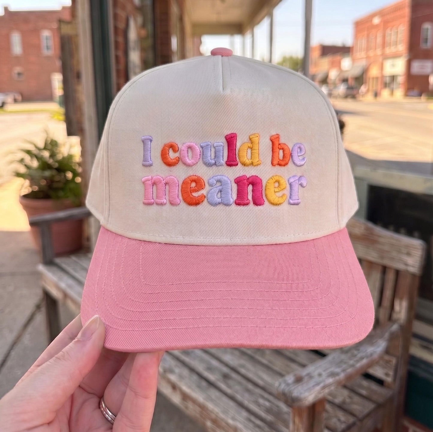 Embroidered cap with colorful 'I Could Be Meaner' text, cream crown with pink brim, displayed in outdoor setting