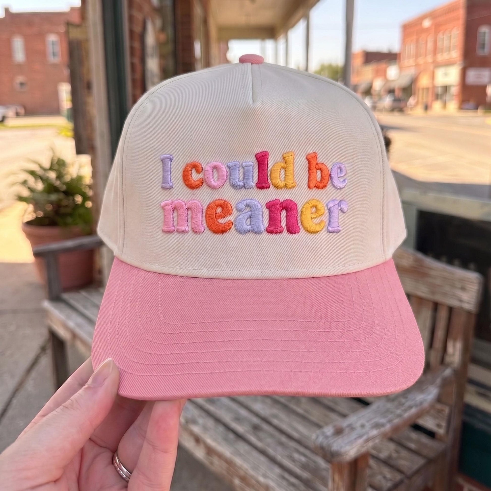 Embroidered cap with colorful 'I Could Be Meaner' text, cream crown with pink brim, displayed in outdoor setting