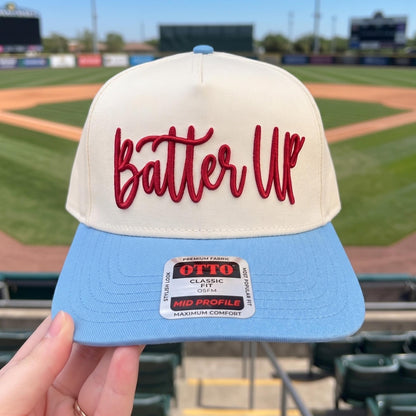 Baseball cap with embroidered 'Batter Up' design in cream and light blue displayed in a stadium setting