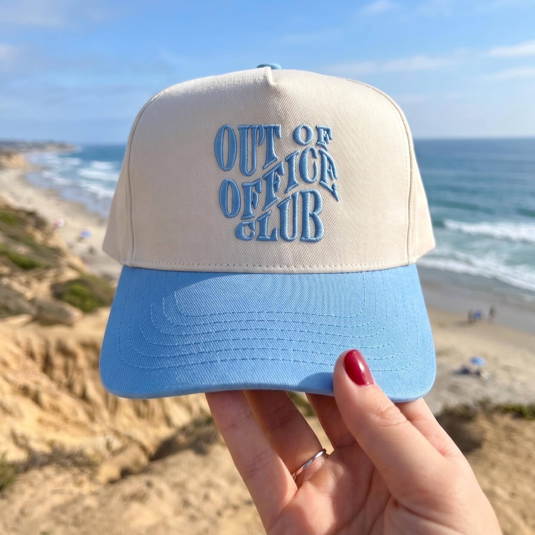 Out of Office Club cap featuring a cream and light blue two-tone design with embroidered logo, held up at a scenic beach