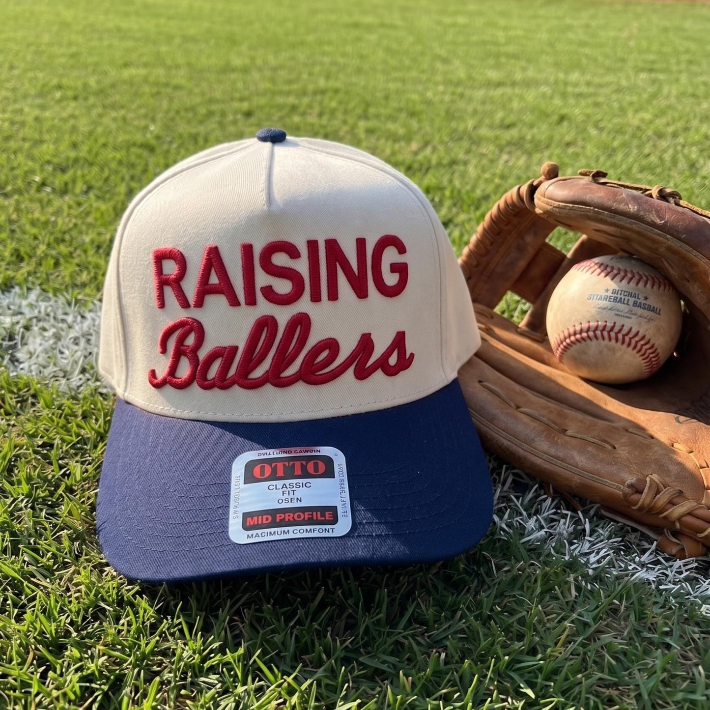 Raising Ballers baseball cap with vintage embroidery in cream and navy displayed with a baseball glove and ball on grass