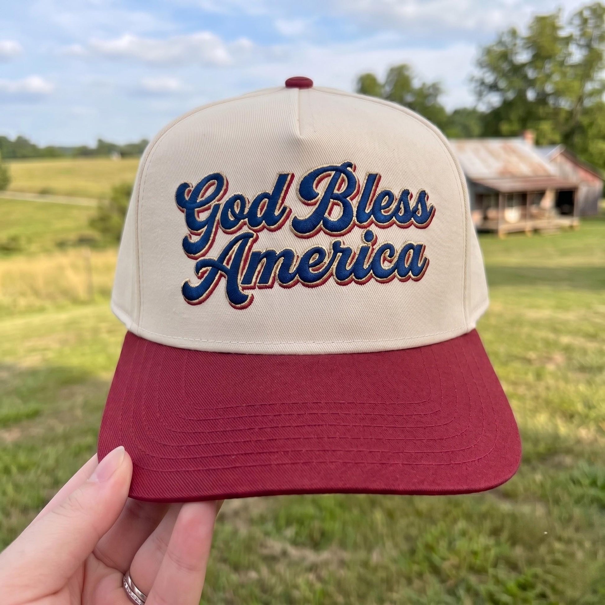 Patriotic trucker hat with 'God Bless America' embroidery in red and beige on a grassy field