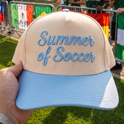 Summer of Soccer baseball cap with beige crown and light blue brim in outdoor setting
