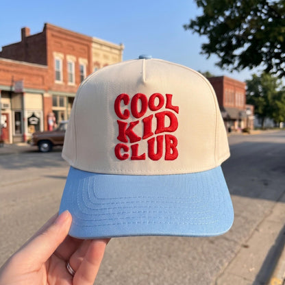 Cool Kid Club embroidered cap with cream and light blue design held on a street backdrop