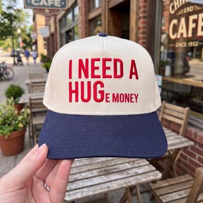 Novelty baseball cap with 'I Need A Huge Money' quote in red embroidery, white crown, navy brim