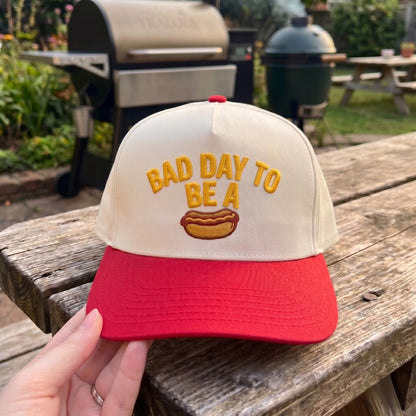 Embroidered snapback hat with 'Bad Day to Be a Hotdog' design in red and cream, displayed outdoors