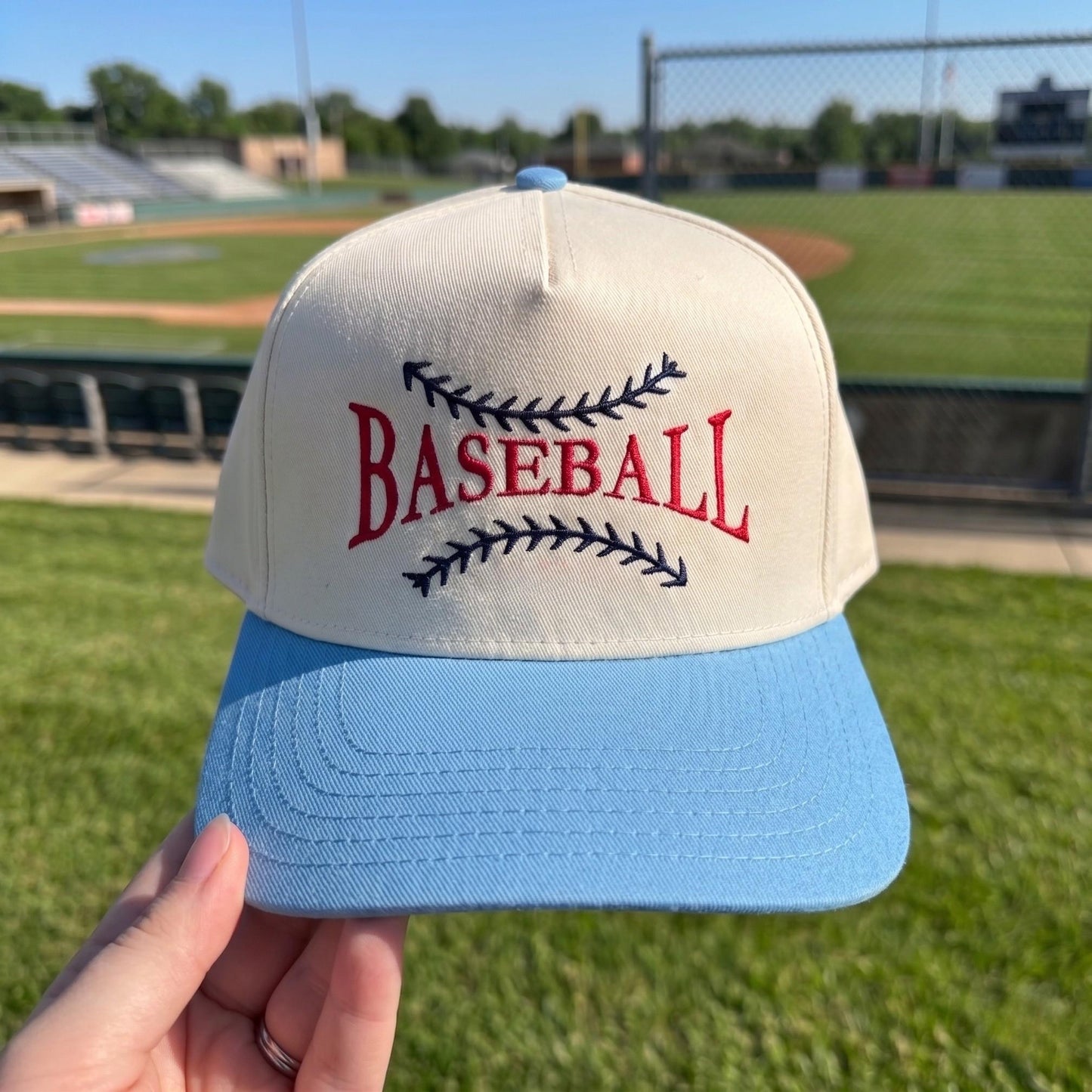 Classic baseball cap with cream crown, light blue brim, and red baseball embroidery at stadium