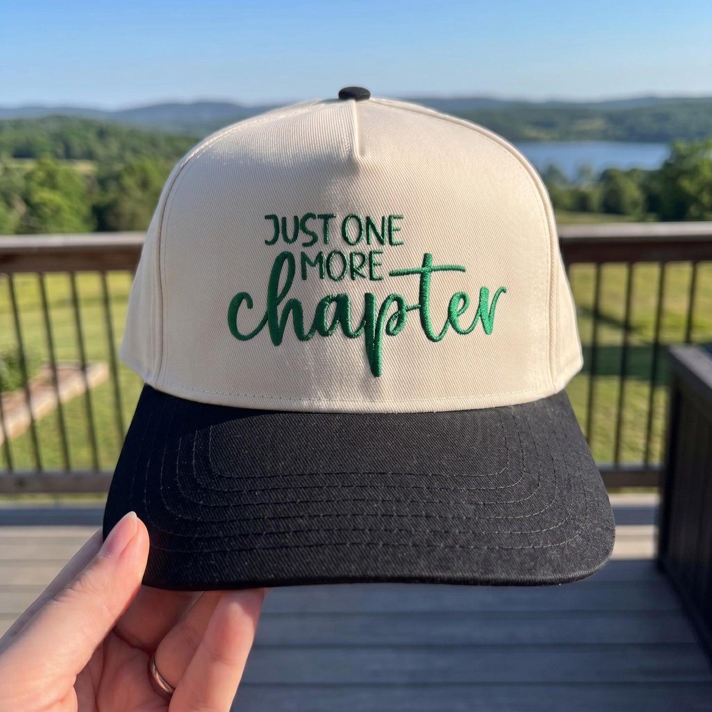 Stylish trucker hat with 'Just One More Chapter' embroidery in green on cream front and black brim set against a scenic