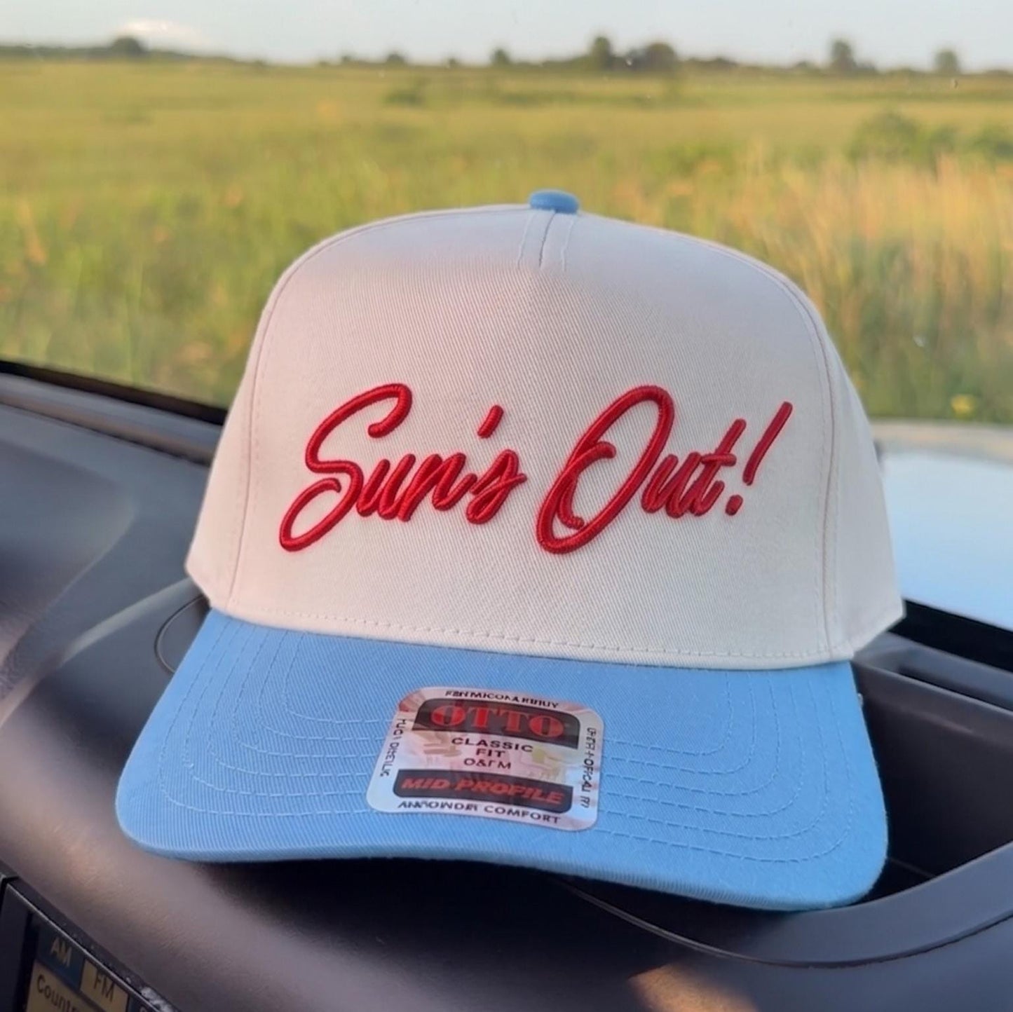 White and blue snapback cap with 'Sun's Out!' embroidery on the dashboard against a green field backdrop