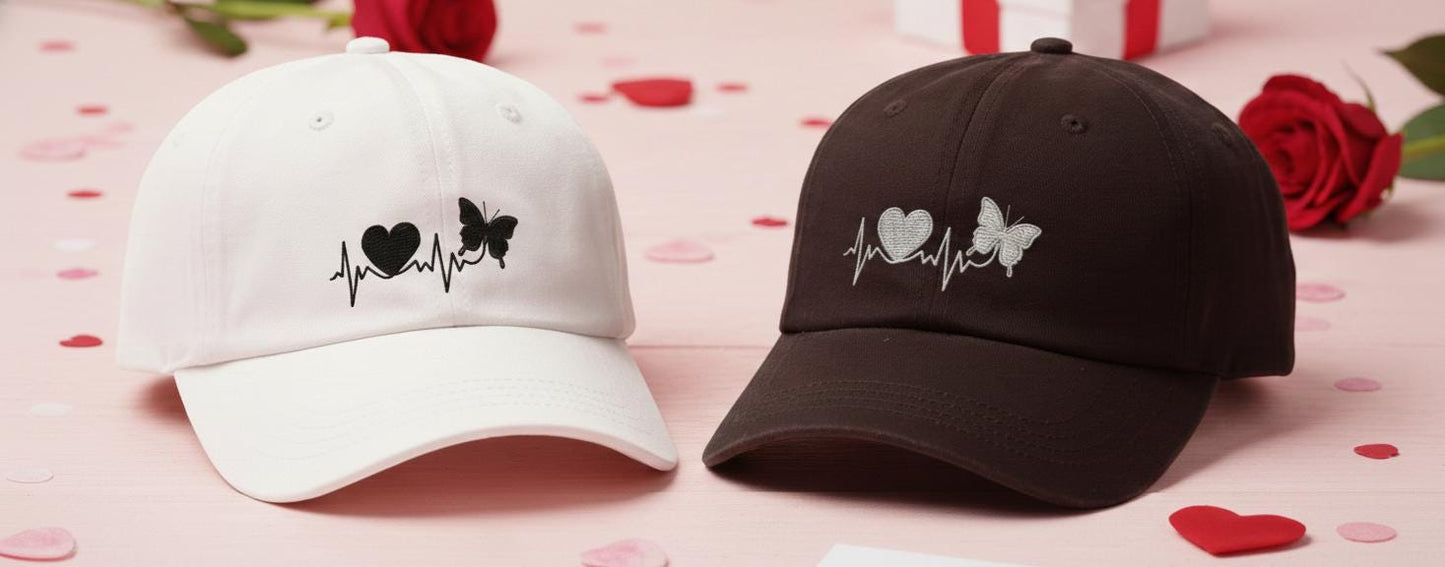 White and black baseball caps with embroidered heartbeat design featuring a heart and butterfly, set on a romantic