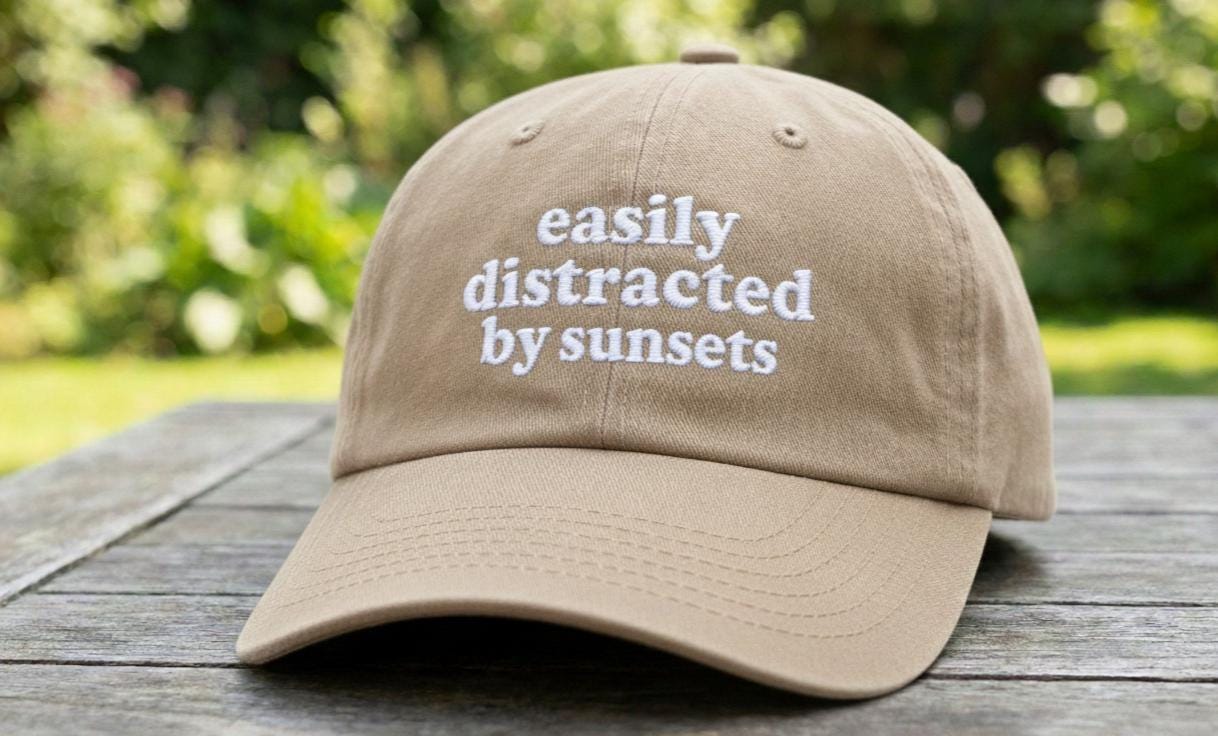 Beige baseball cap with 'easily distracted by sunsets' white embroidery on a wooden picnic table