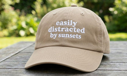 Beige baseball cap with 'easily distracted by sunsets' white embroidery on a wooden picnic table