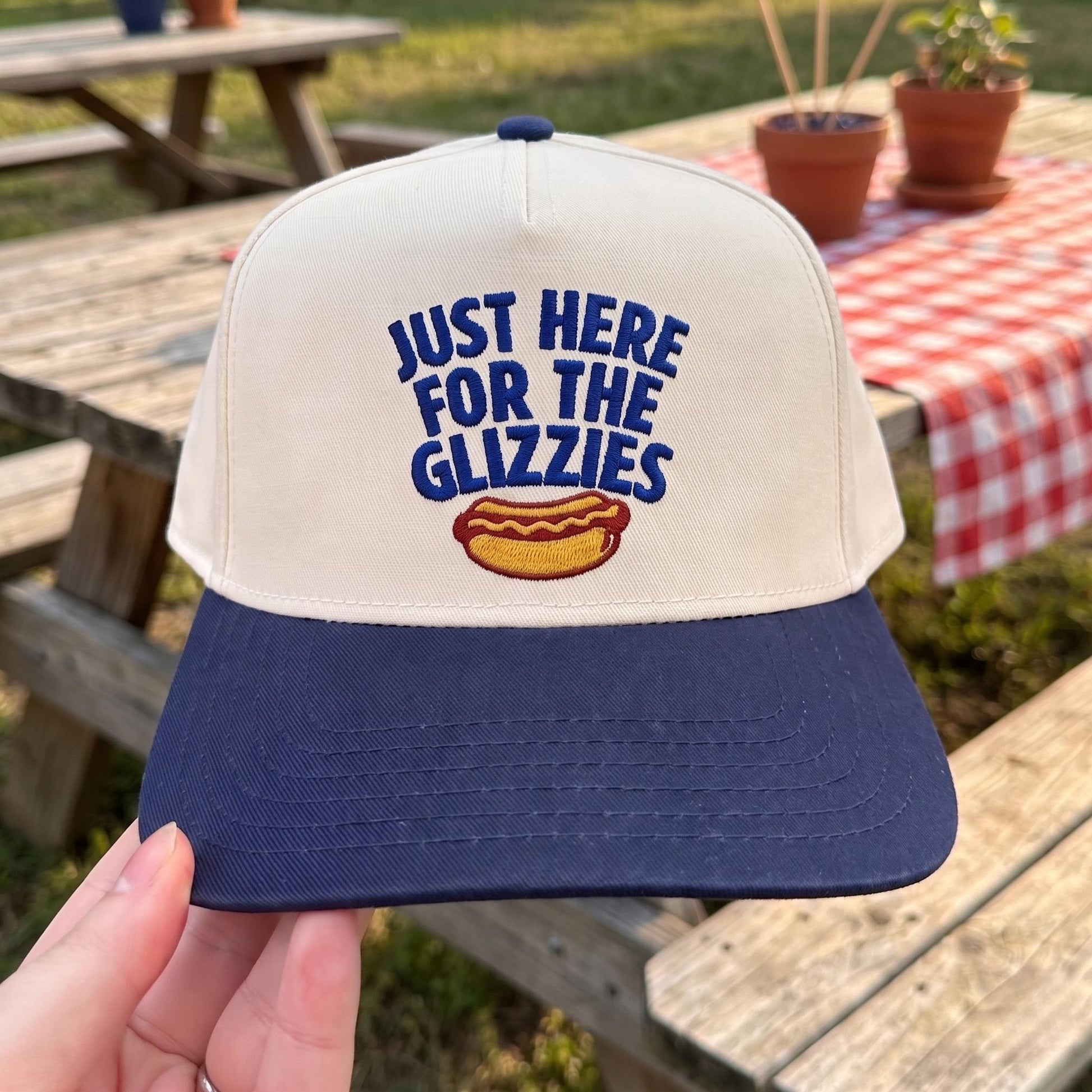 Trendy baseball cap with 'Just Here for the Glizzies' slogan in beige and navy, displayed outdoors on a picnic table