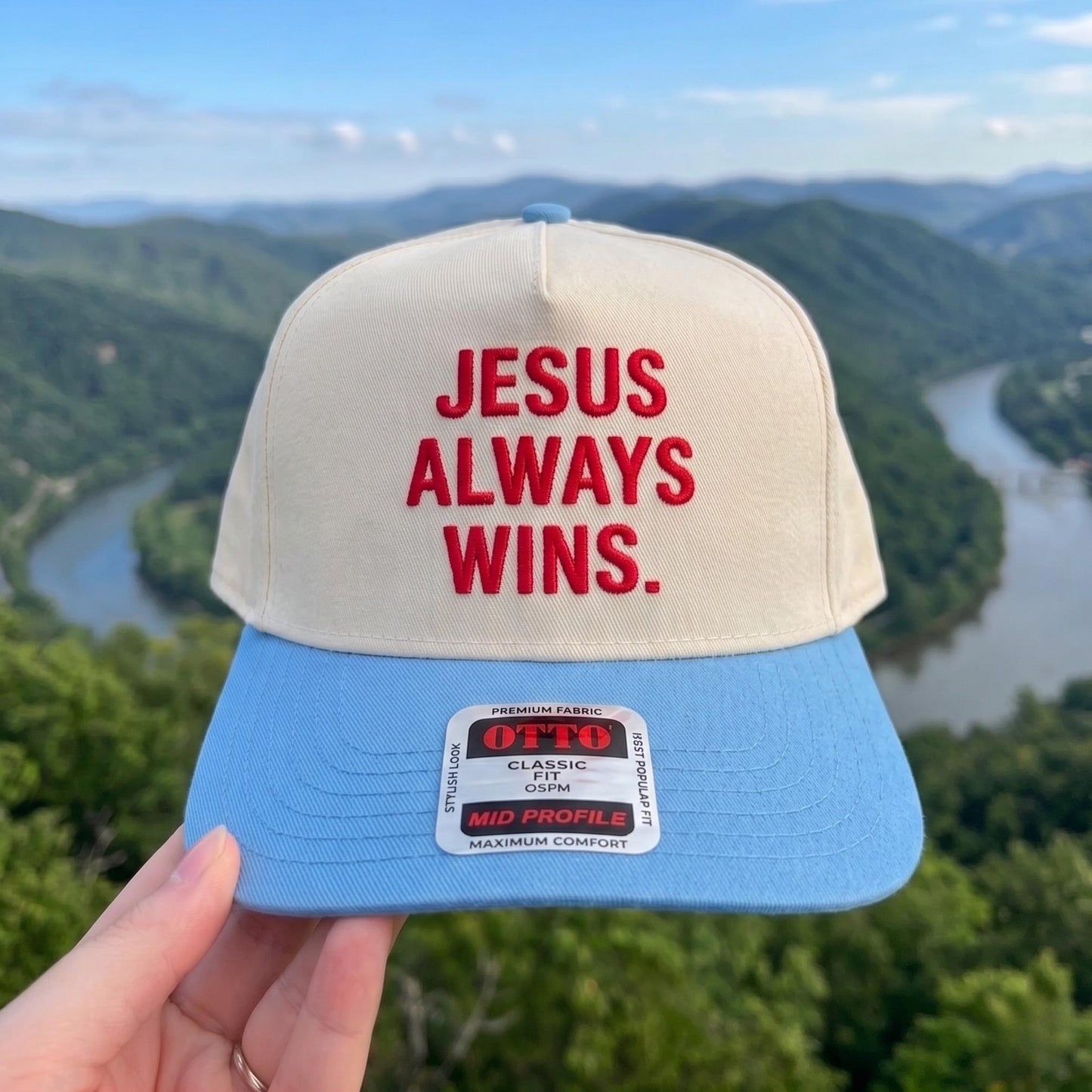 Jesus Always Wins embroidered baseball cap with beige top and blue bill, scenic background