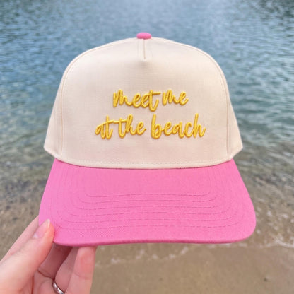 Trendy beach cap with beige crown and pink visor featuring 'Meet Me at the Beach' embroidered in yellow, held at the ocean