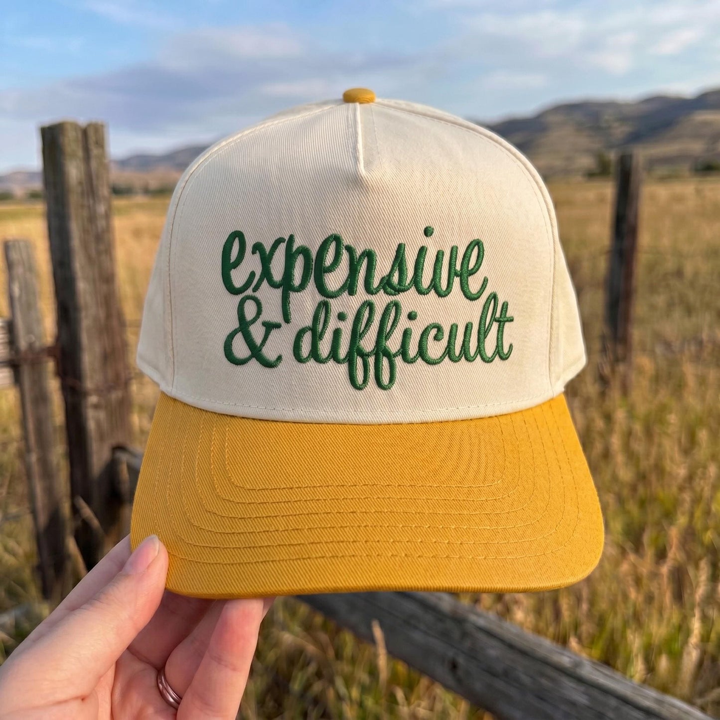 Expensive & Difficult' embroidered trucker hat with yellow brim and white panel in outdoor setting