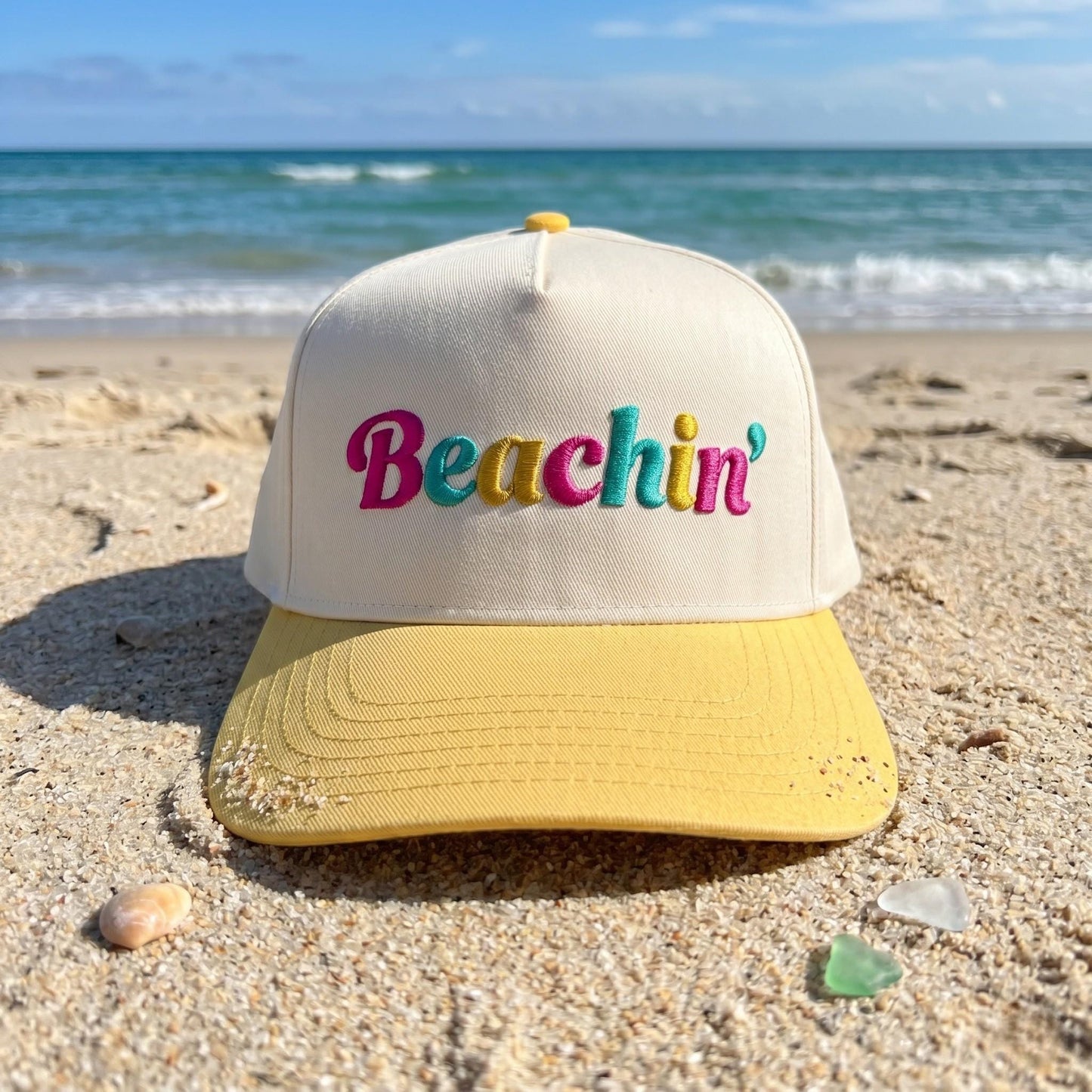 Embroidered baseball cap with 'Beachin'' in colorful pink, teal, and yellow on cream crown and yellow brim, displayed on
