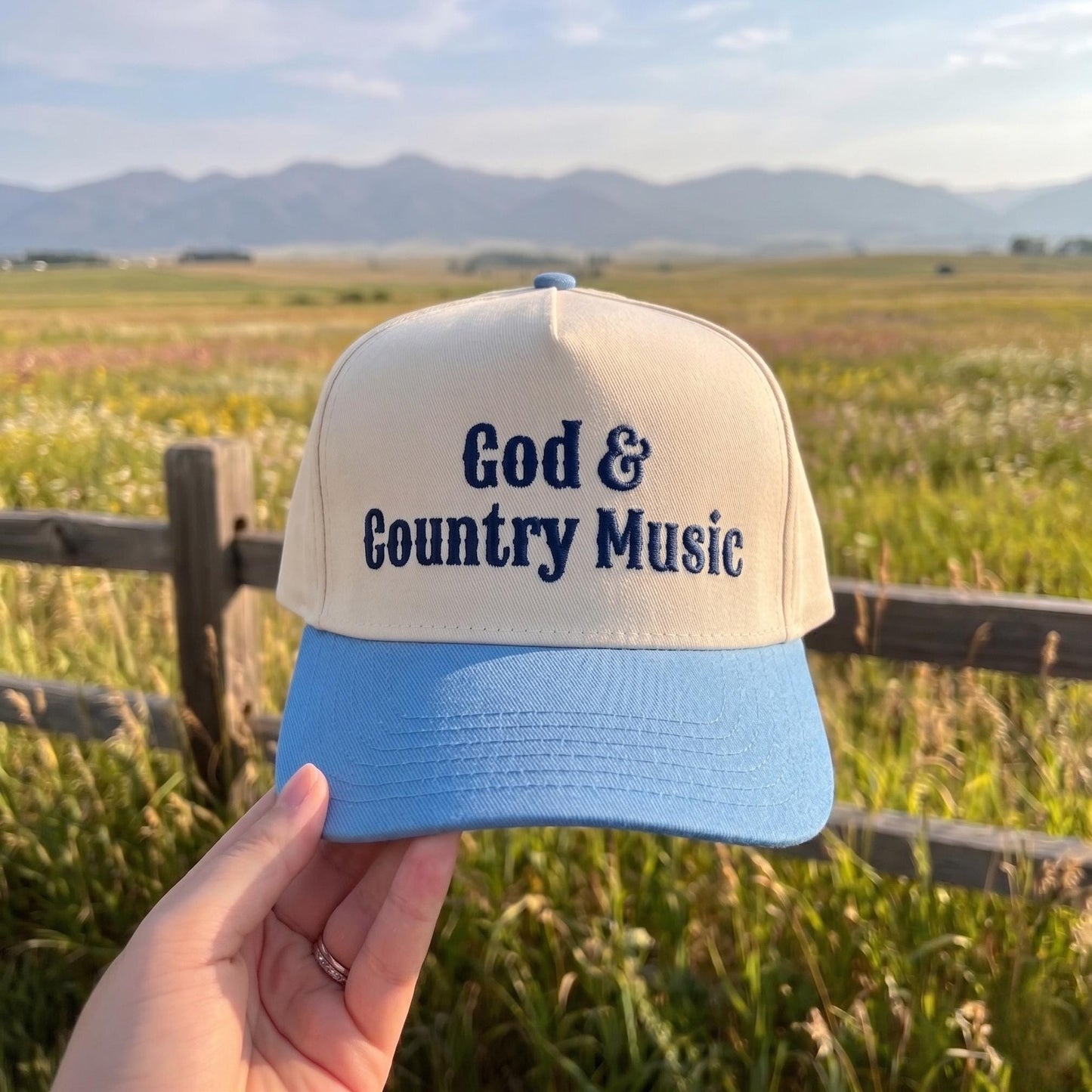 Cream and blue baseball cap with 'God & Country Music' embroidery held against a scenic field background