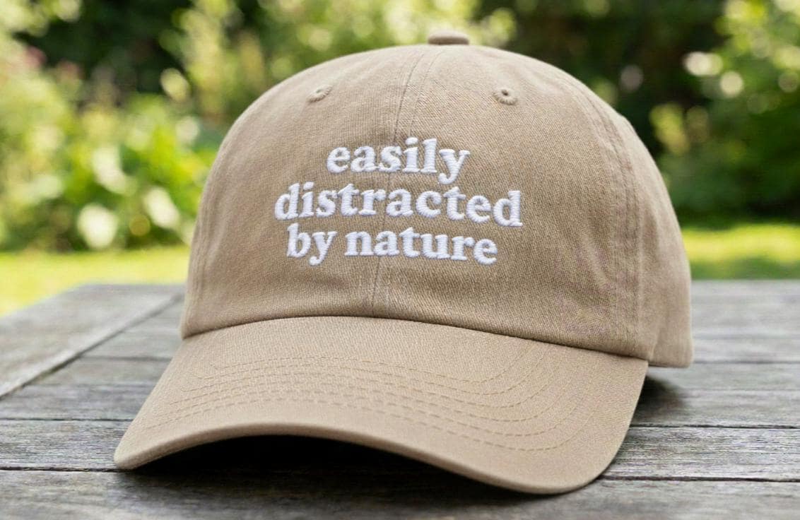 Beige baseball cap with white embroidered text 'easily distracted by nature' on a wooden table outdoors