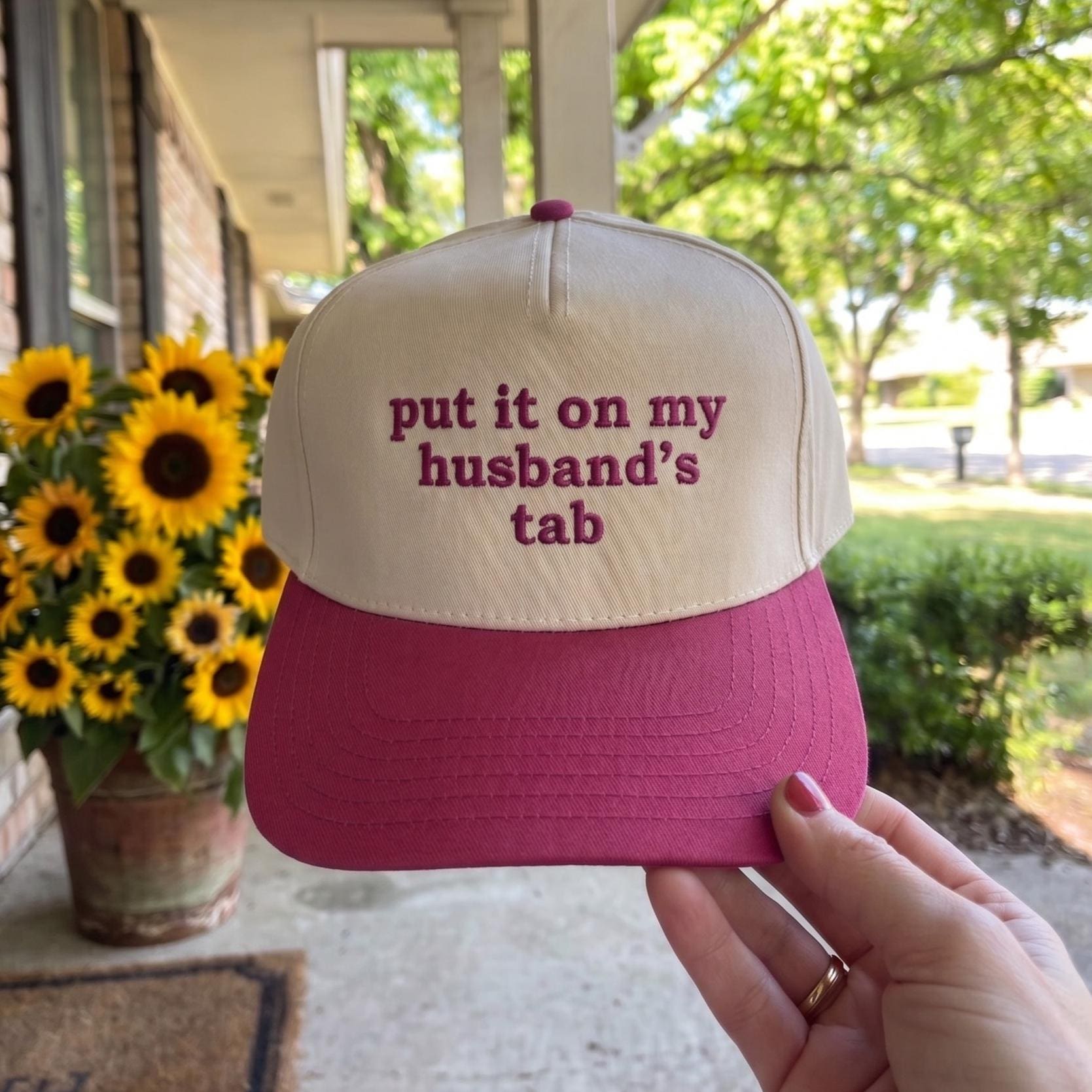 Fashionable women's cap in pink and white with humorous quote 'put it on my husband's tab' embroidery held outdoors