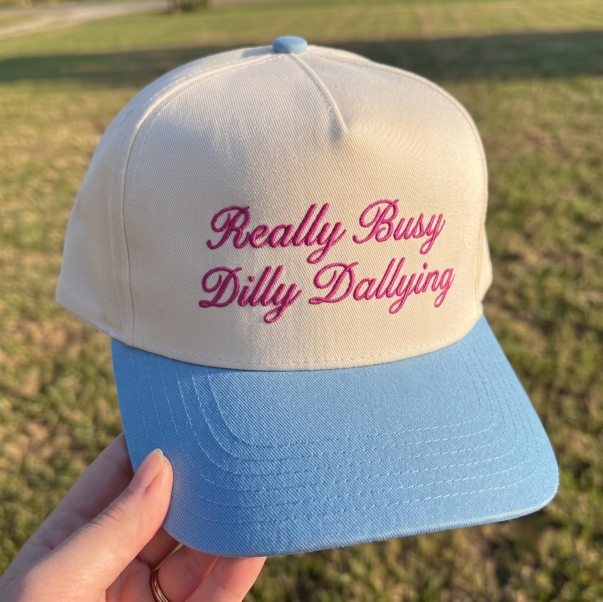 Embroidered baseball hat with 'Really Busy Dilly Dallying' in pink text on cream with light blue brim